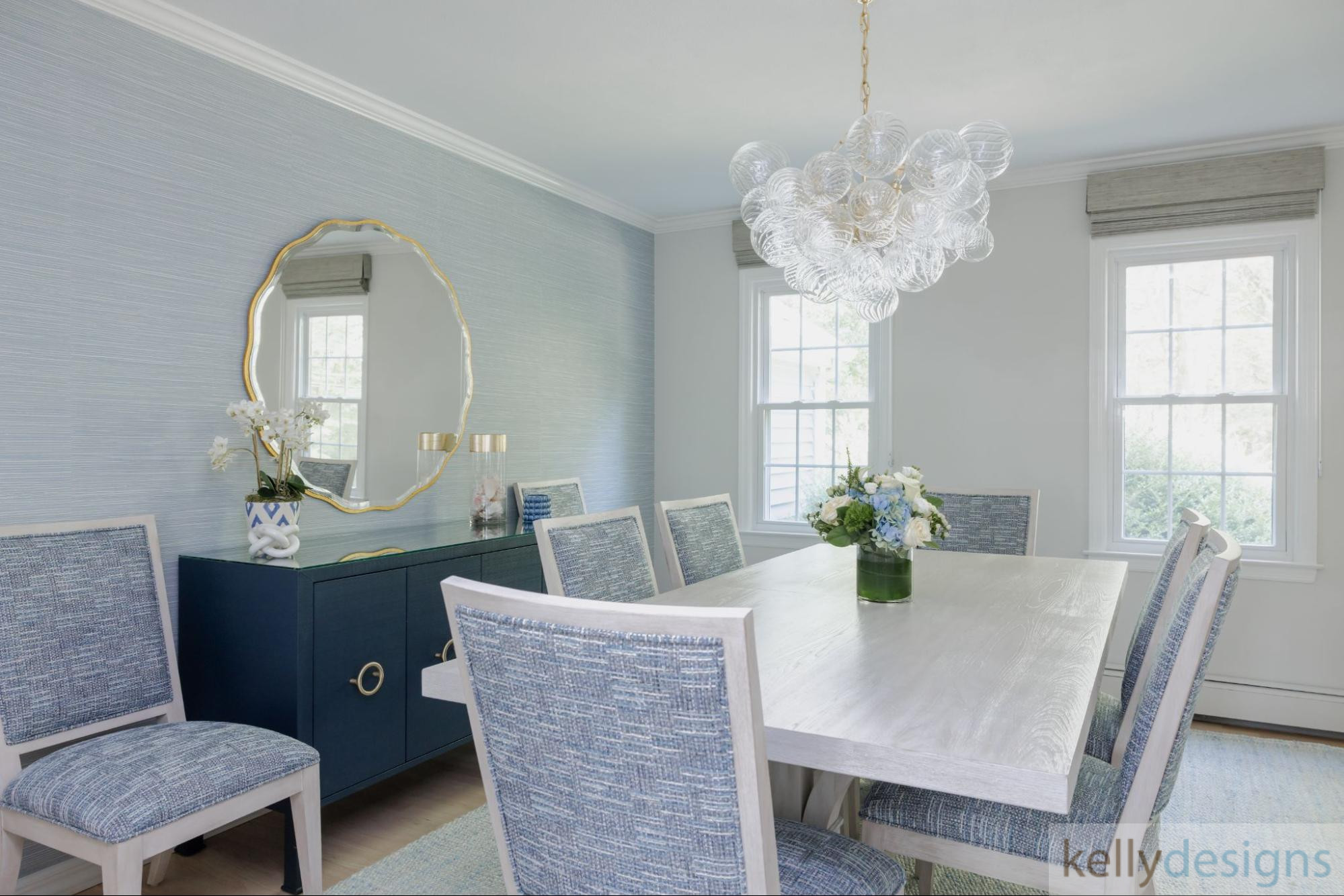 A sophisticated dining room featuring a long wooden table with gray upholstered chairs, a large white crystal statement chandelier, and a blue accent wall with a large window.