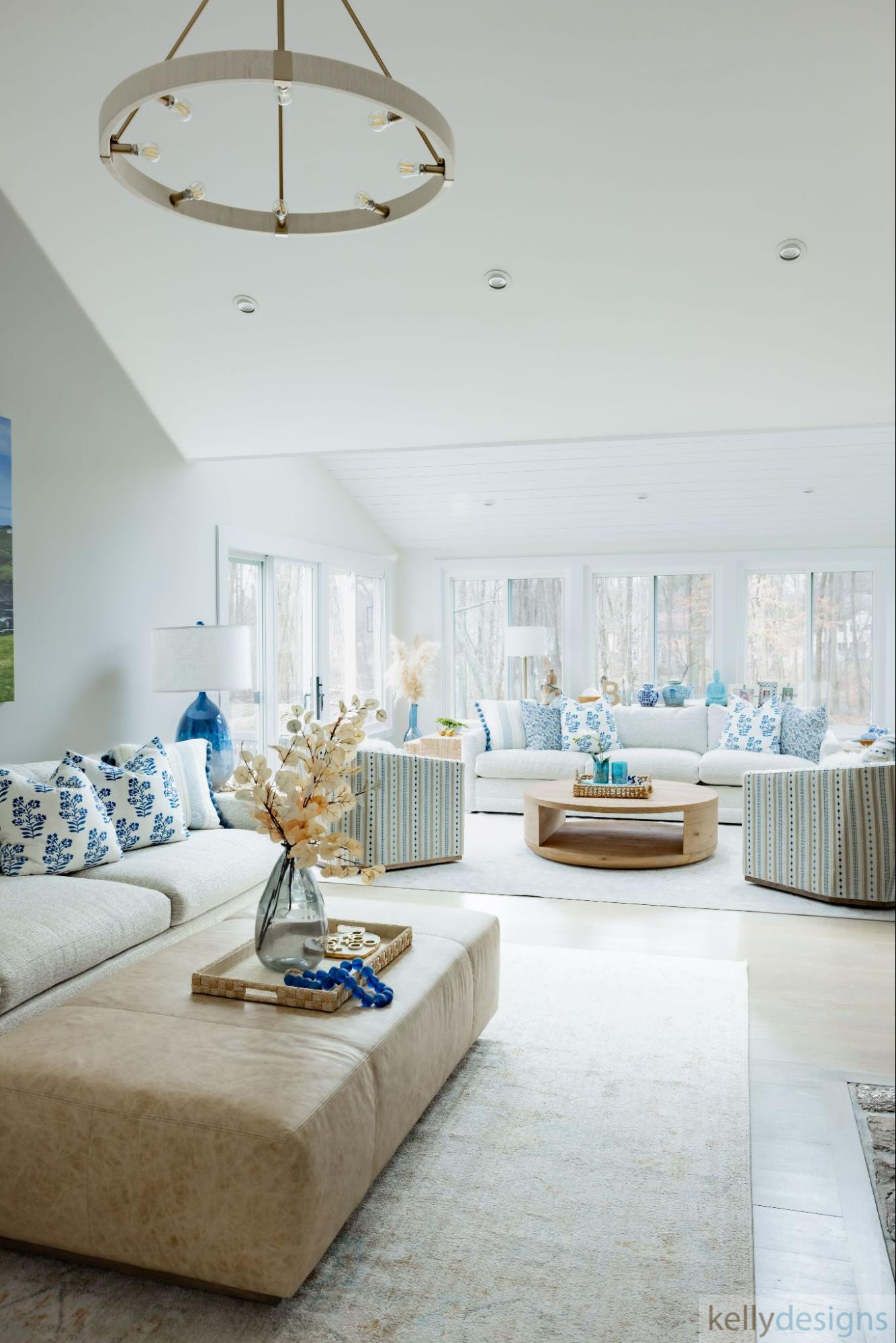 A light-filled living room featuring a white sectional sofa, light blue accent pillows, a marble-top coffee table, and large windows with a view of a green landscape.