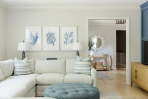A bright living room featuring a white sectional sofa with blue patterned pillows, a round blue tufted ottoman used as a coffee table, and three pieces of blue botanical wall art.
