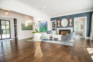 A bright, modern living room with a neutral sectional sofa, a round glass-top coffee table with a gold base, and a large window providing natural light, looking through to a dining area.