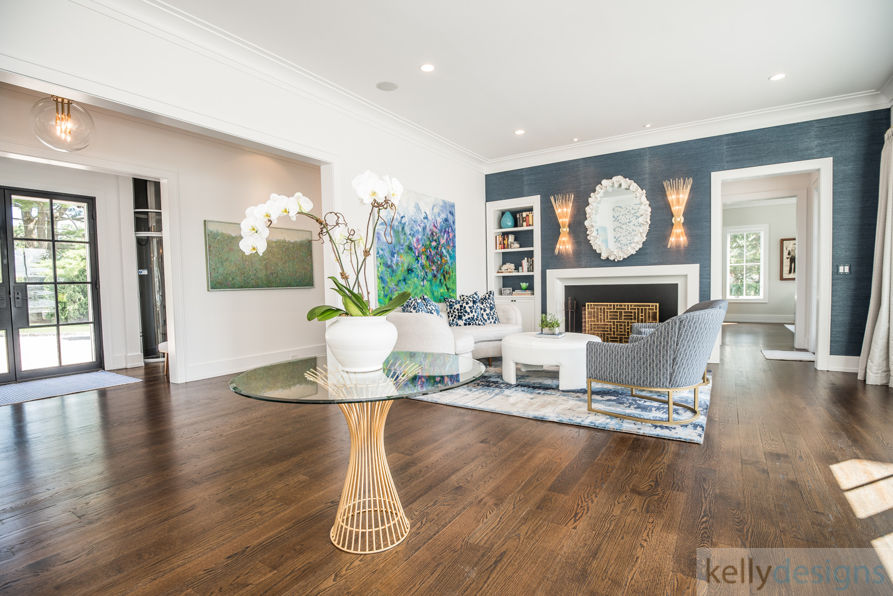 A bright, modern living room with a neutral sectional sofa, a round glass-top coffee table with a gold base, and a large window providing natural light, looking through to a dining area.