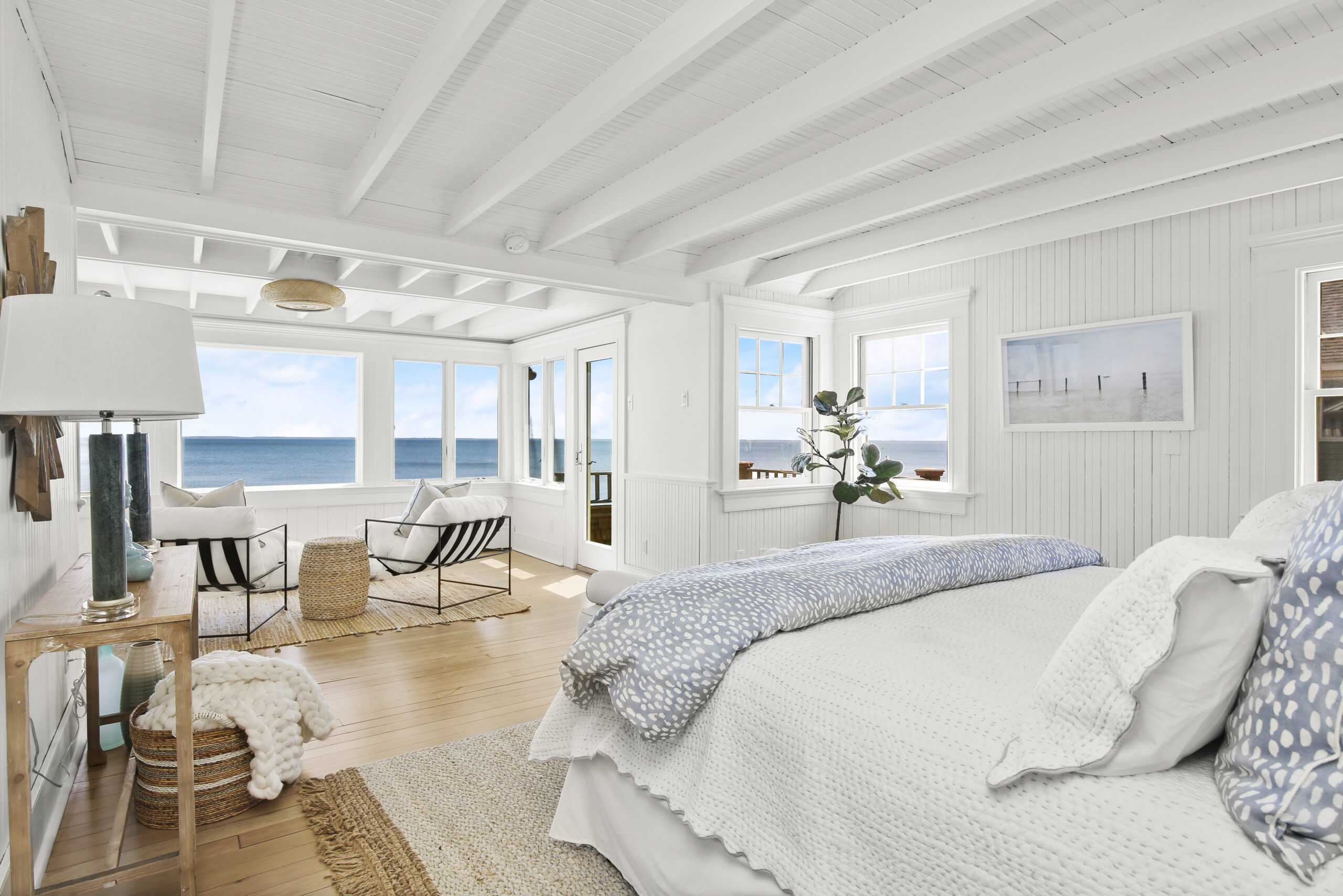 Coastal-style master bedroom sitting area with a white chaise lounge, natural wood beamed ceiling, and large windows with an ocean view.