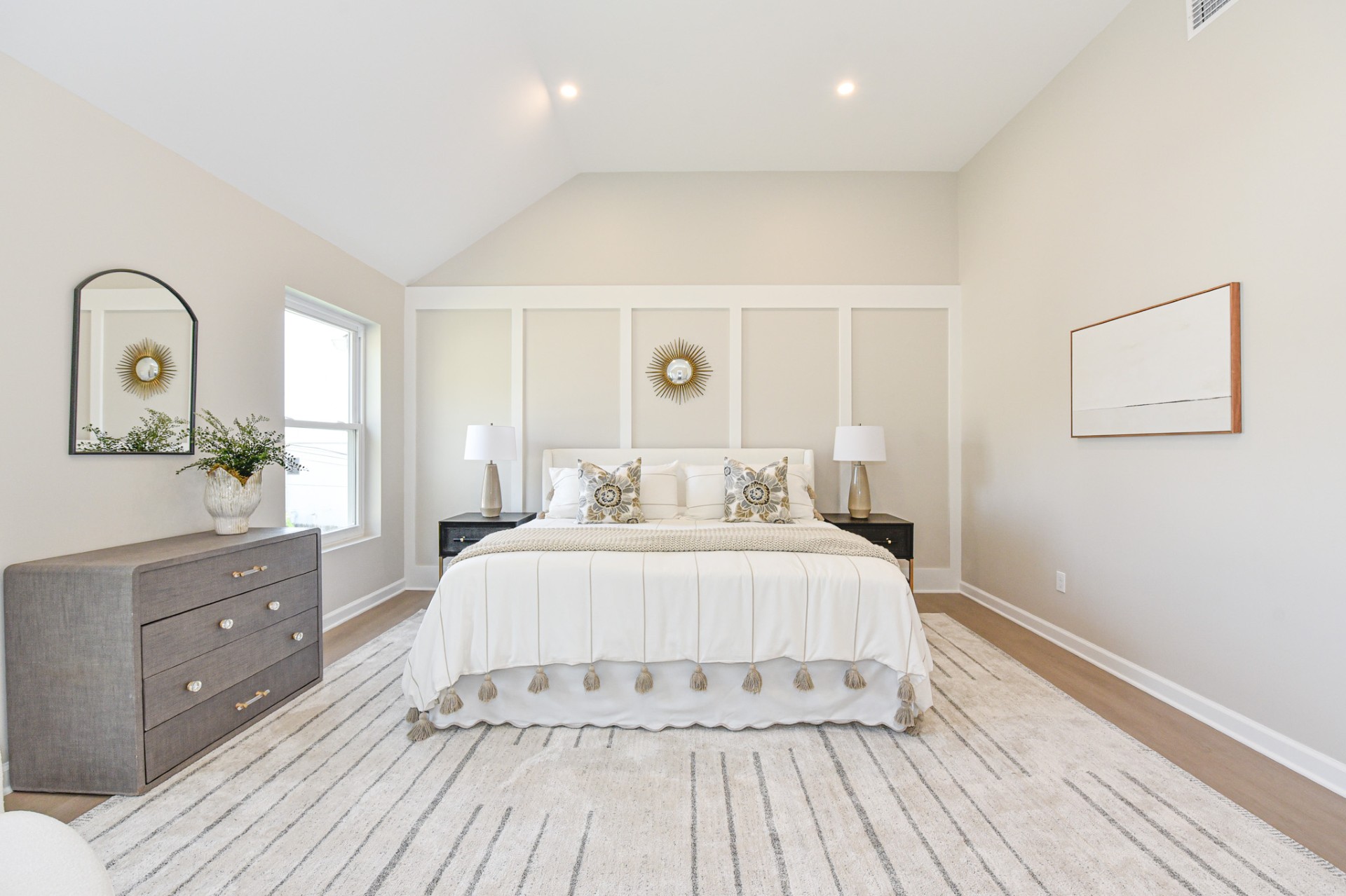 Bright modern master suite with a neutral color palette, featuring a white upholstered bed, a vaulted ceiling, and large windows overlooking greenery.