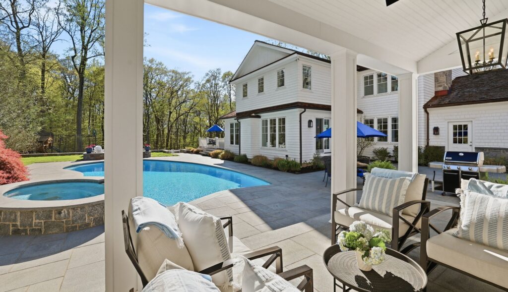 A luxurious white colonial-style home in Westport, CT, with a large in-ground pool and a covered patio featuring comfortable outdoor seating and a modern grill.
