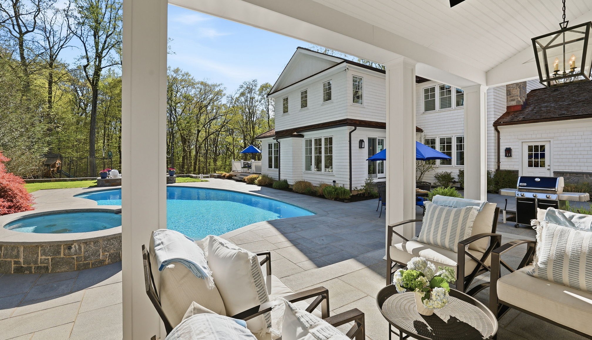 A luxurious white colonial-style home in Westport, CT, with a large in-ground pool and a covered patio featuring comfortable outdoor seating and a modern grill.